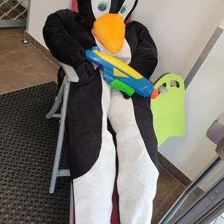 Pinguin-Kost&uuml;m sitzt in einem Stuhl und h&auml;lt eine Wasserpistole in der Hand. Heller Hintergrund mit mattem Boden.