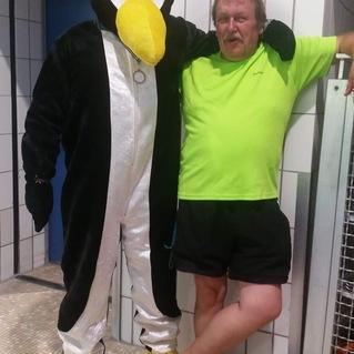 Eine Person in einem Pinguinkost&uuml;m steht neben einem Mann in einem gr&uuml;nen Shirt und schwarzer Badehose in einem Schwimmbad.