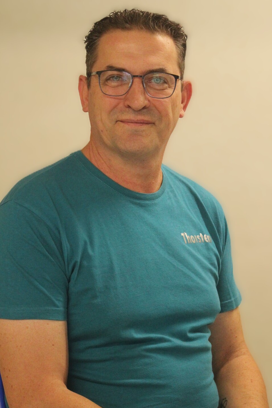 Mann mit Brille sitzt l&auml;chelnd vor einem hellen Hintergrund, tr&auml;gt ein t&uuml;rkisfarbenes T-Shirt.