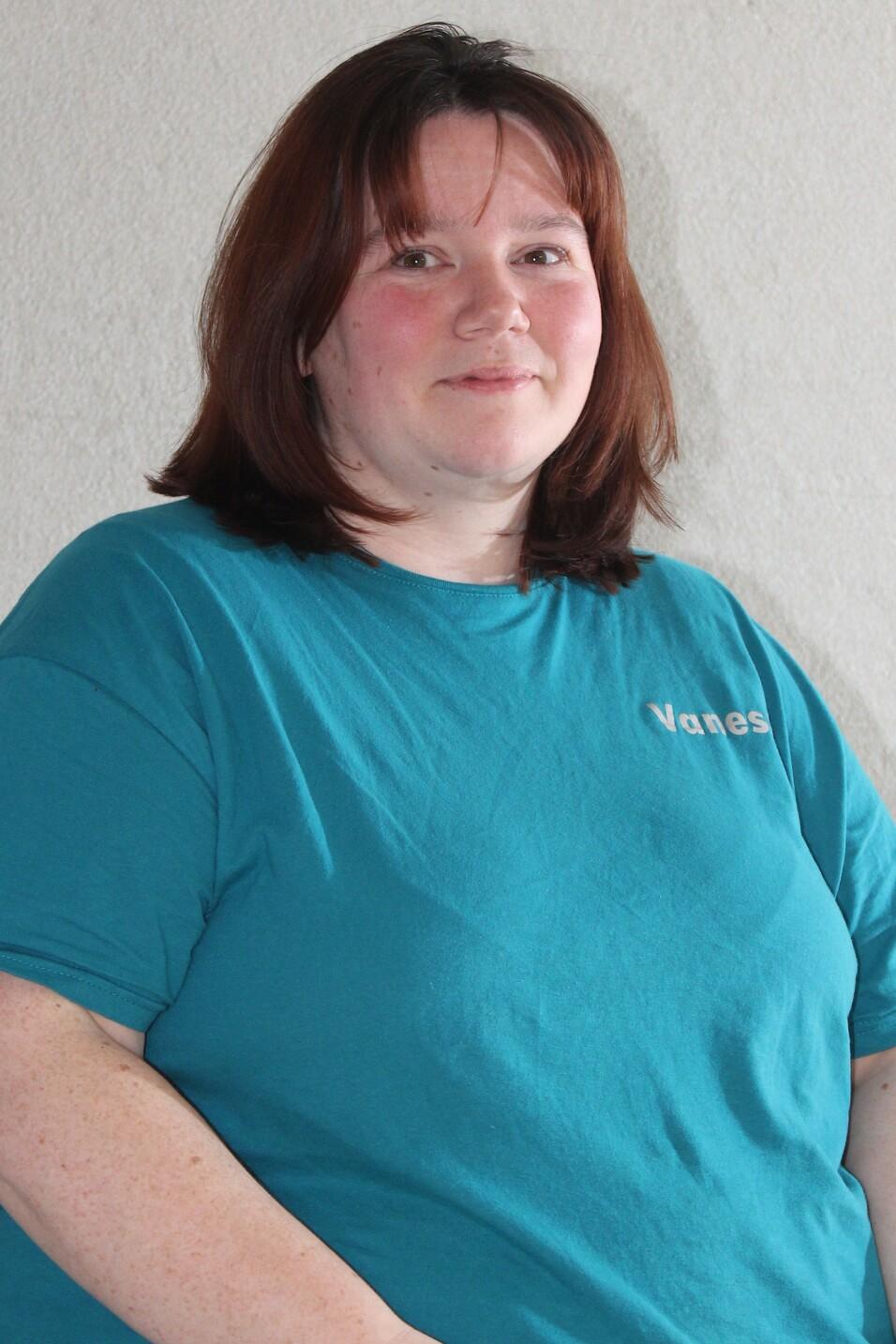 Frau mit schulterlangen, braunen Haaren, tr&auml;gt ein t&uuml;rkisfarbenes T-Shirt, sitzt auf einem Stuhl vor einer grauen Wand.