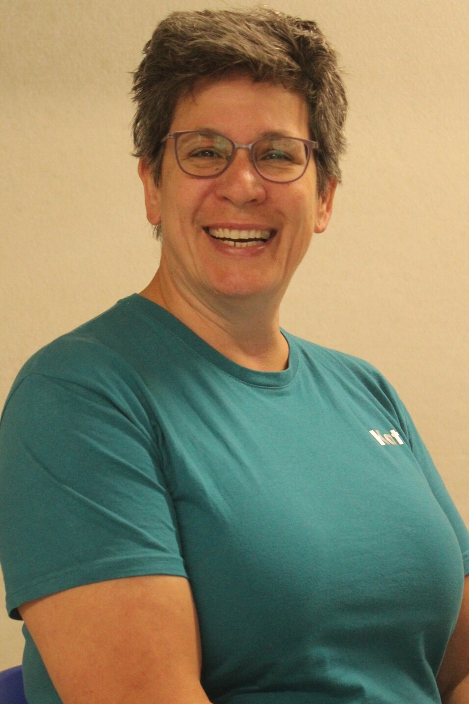 Lachende Frau mit kurzem grauen Haar, Brille und t&uuml;rkisfarbenem T-Shirt, sitzend vor heller Wand.