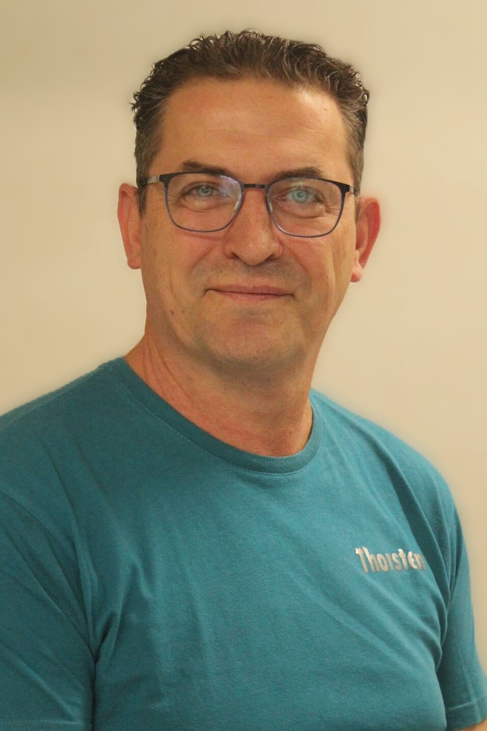 Mann mit Brille sitzt l&auml;chelnd vor einem hellen Hintergrund, tr&auml;gt ein t&uuml;rkisfarbenes T-Shirt.