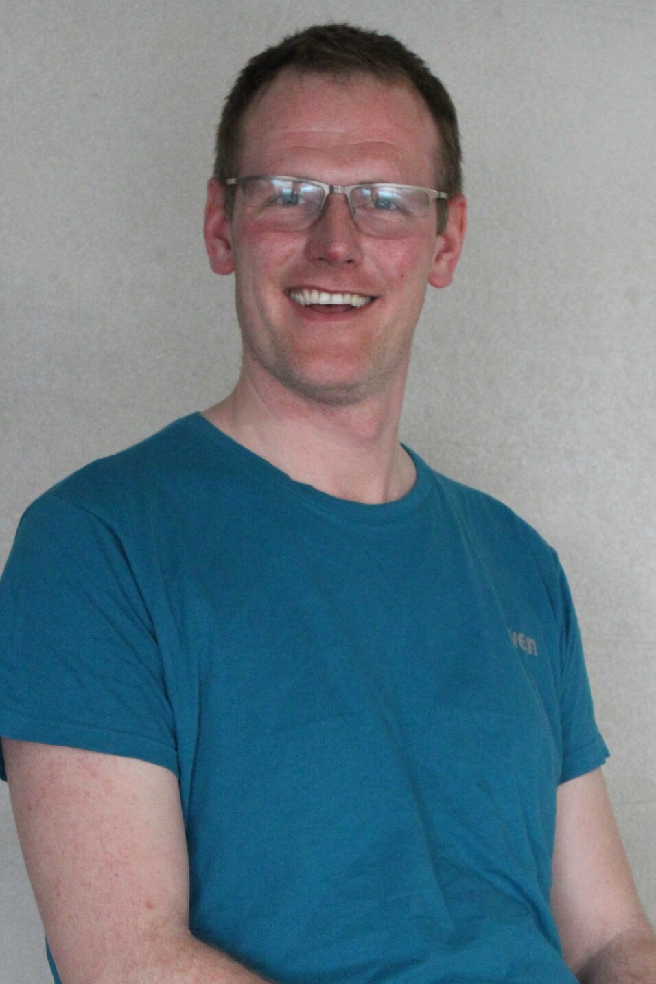 L&auml;chelnder Mann mit Brille in blauem T-Shirt sitzt vor einer hellen Wand.