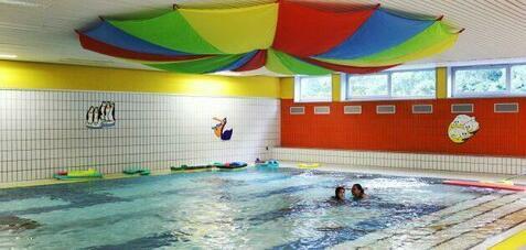 Blick auf ein Hallenbad mit buntem Deckenvorhang, Wasserspielzeug und schwimmenden Kindern im Wasser.