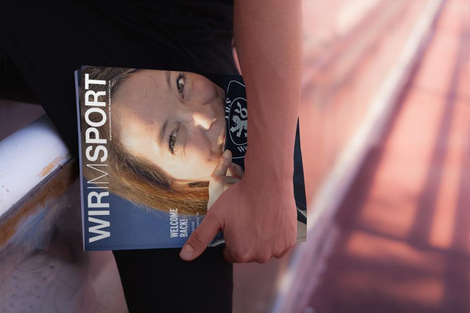 Nahaufnahme einer Person, die ein Magazin mit dem Titel &bdquo;WIR IM SPORT&ldquo; in der Hand h&auml;lt, im Hintergrund eine Sportfl&auml;che.