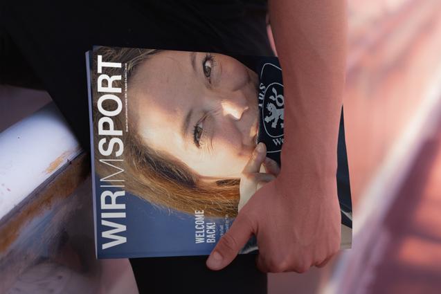 Nahaufnahme einer Person, die ein Magazin mit dem Titel &bdquo;WIR IM SPORT&ldquo; in der Hand h&auml;lt, im Hintergrund eine Sportfl&auml;che.